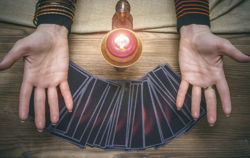 在木質桌面上，一名占卜師張開雙手展示前方呈扇形排列的塔羅牌，中間點燃著一顆紅色蠟燭，營造出神祕且充滿靈性指引的氛圍。