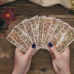 在木質桌面上，一名占卜師雙手拿著呈扇形展開的塔羅牌。周圍佈置了紅花裝飾的金色蠟燭、天然原石、金色葉片裝飾以及占卜用的小道具，營造出神祕且莊重的儀式感