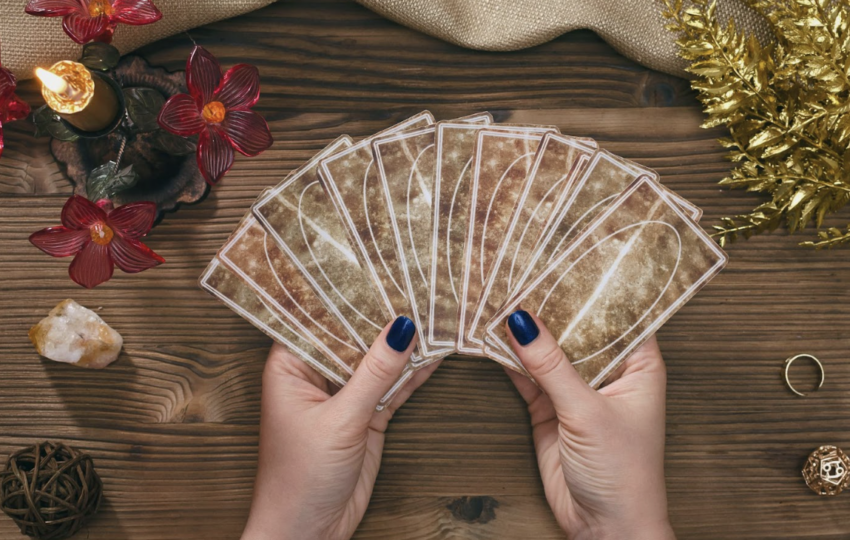 在木質桌面上，一名占卜師雙手拿著呈扇形展開的塔羅牌。周圍佈置了紅花裝飾的金色蠟燭、天然原石、金色葉片裝飾以及占卜用的小道具，營造出神祕且莊重的儀式感