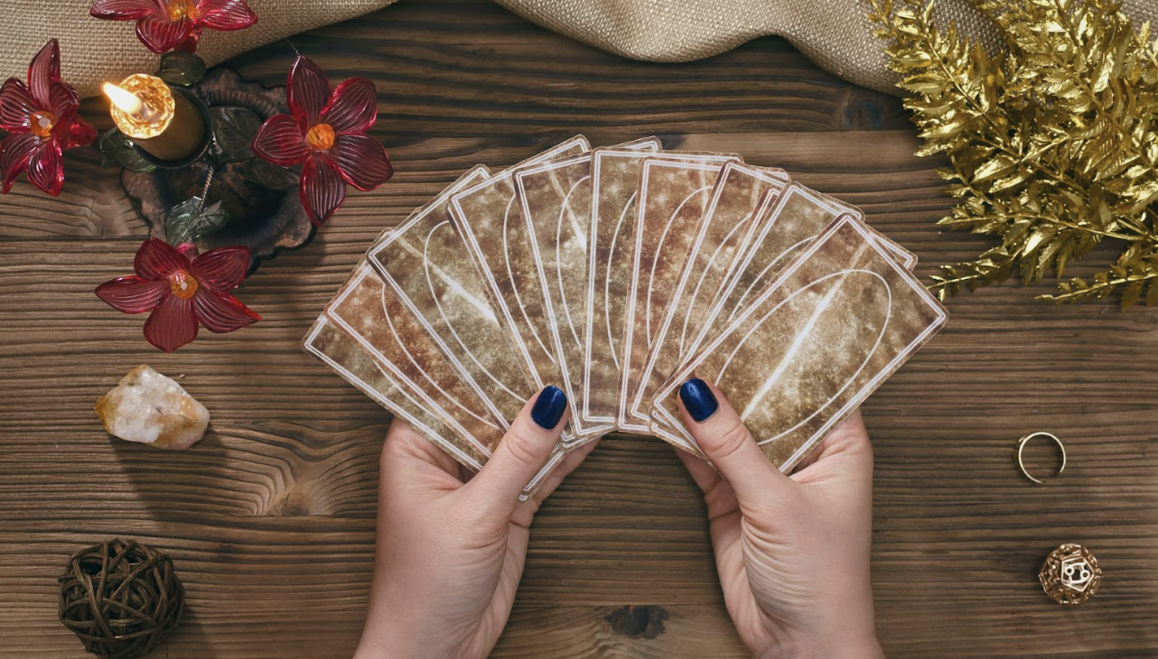 在木質桌面上，一名占卜師雙手拿著呈扇形展開的塔羅牌。周圍佈置了紅花裝飾的金色蠟燭、天然原石、金色葉片裝飾以及占卜用的小道具，營造出神祕且莊重的儀式感