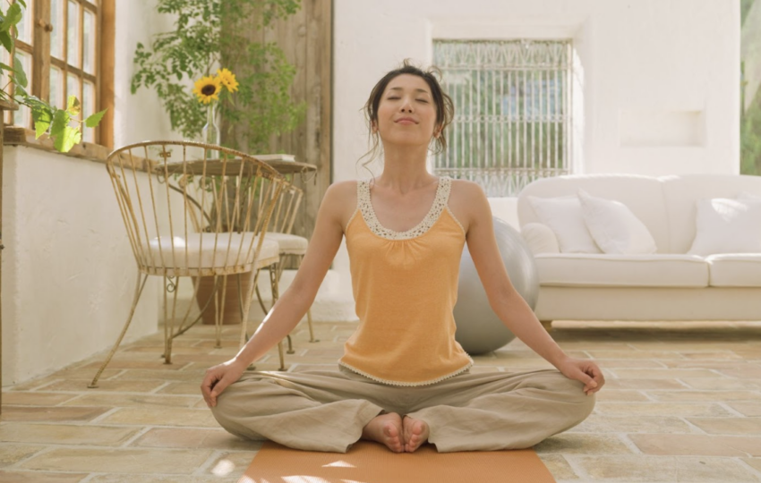 一位女性在充滿自然光的溫馨客廳地板上盤腿冥想，背景有白色沙發與綠意植栽。
