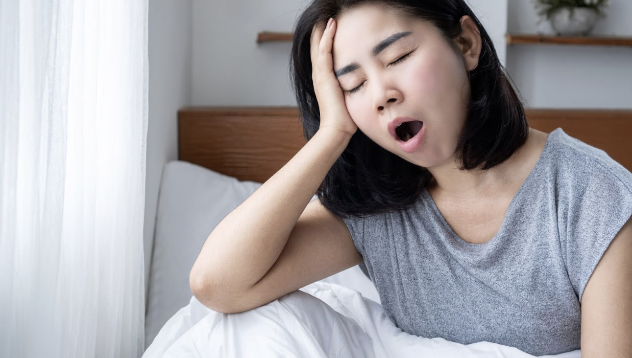 一位疲憊的女性在窗邊床上打哈欠並扶著額頭，暗示睡眠不足或清晨光線過亮的困擾