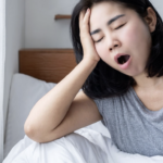 一位疲憊的女性在窗邊床上打哈欠並扶著額頭，暗示睡眠不足或清晨光線過亮的困擾