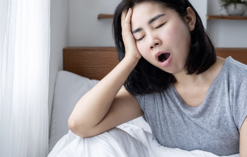 一位疲憊的女性在窗邊床上打哈欠並扶著額頭，暗示睡眠不足或清晨光線過亮的困擾