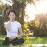 陽光灑落在綠色草地上，女性閉眼進行調息法冥想，背景是逆光下的自然樹影。