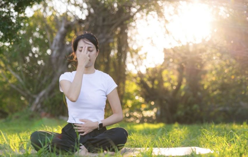 陽光灑落在綠色草地上，女性閉眼進行調息法冥想，背景是逆光下的自然樹影。