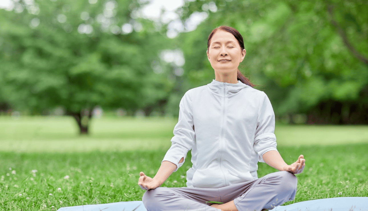 中年女性在戶外草地上進行盤腿冥想，閉目感受自然氣息，展現身心靈合一的放鬆狀態