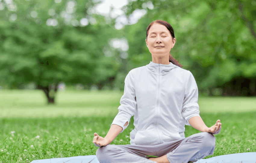中年女性在戶外草地上進行盤腿冥想，閉目感受自然氣息，展現身心靈合一的放鬆狀態