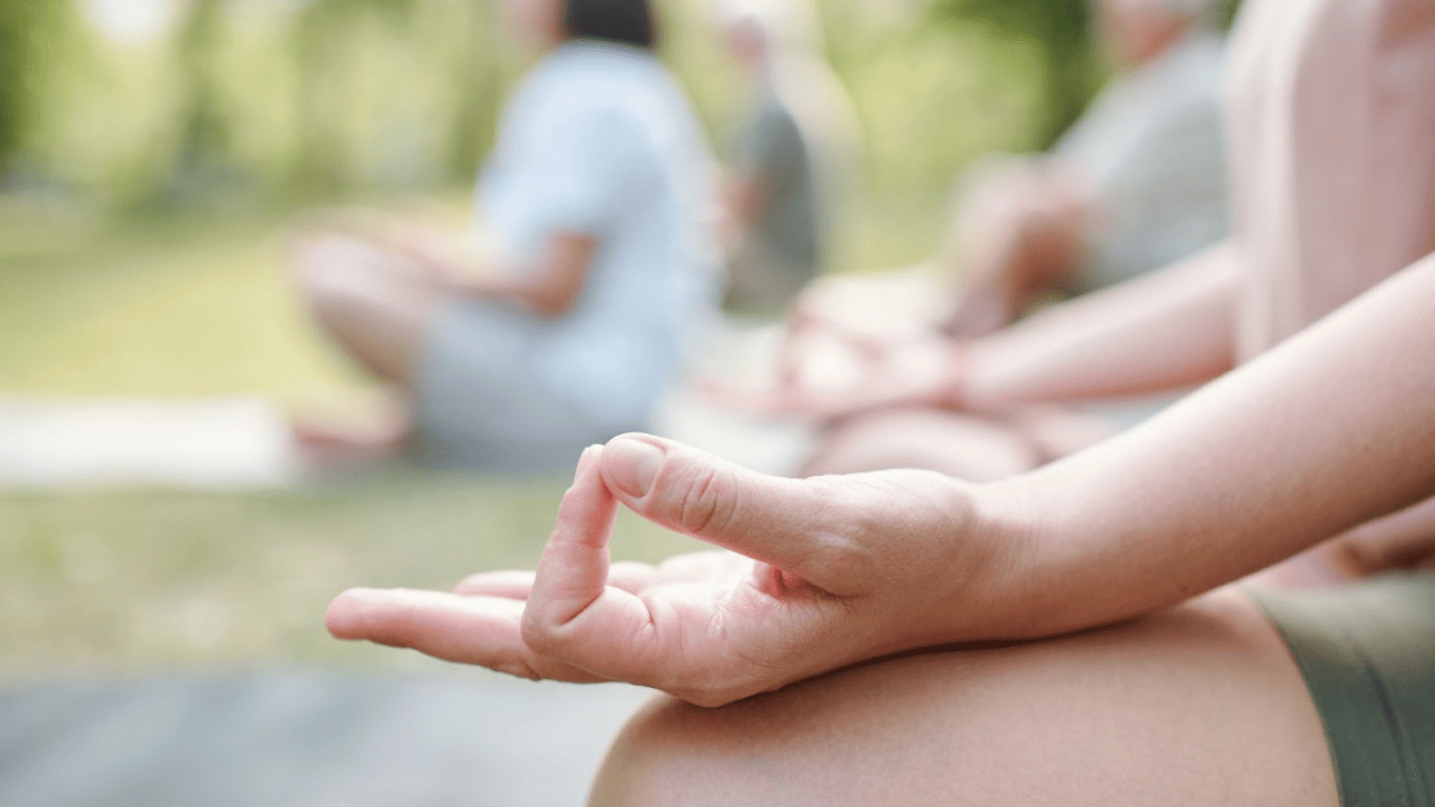 團體瑜伽課程特寫：練習者手部輕放在膝蓋上，手指結印，遠處是共同練習的學員模糊身影