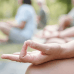 團體瑜伽課程特寫：練習者手部輕放在膝蓋上，手指結印，遠處是共同練習的學員模糊身影