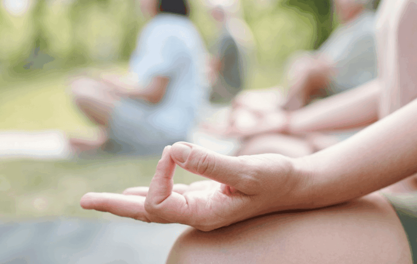 團體瑜伽課程特寫：練習者手部輕放在膝蓋上，手指結印，遠處是共同練習的學員模糊身影