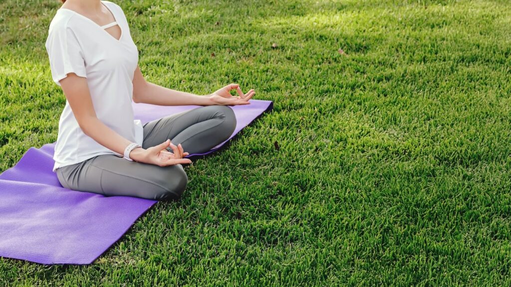 一名女性坐在紫色瑜珈墊上於草地進行盤腿冥想的側影，雙手結印，專注於戶外的靜心練習
