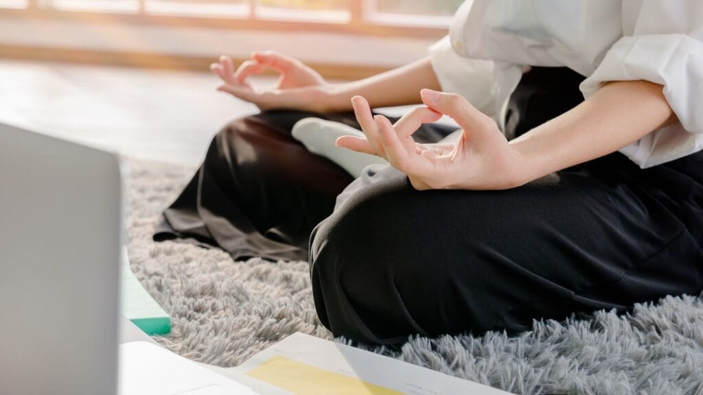  在居家空間的灰色毛絨地毯上進行室內冥想，身旁擺放著筆電與文件，展現忙碌生活中的平靜。