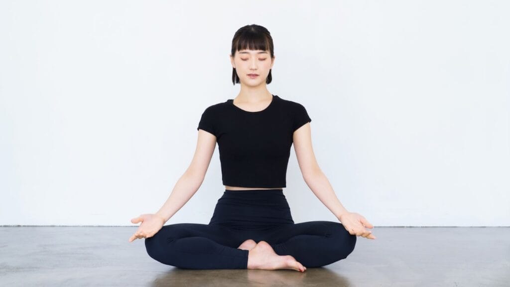  一位年輕女性在純白背景前的平整地面上，雙腿盤坐練習冥想，雙手自然垂放於膝蓋併呈手印狀，表情平靜祥和。