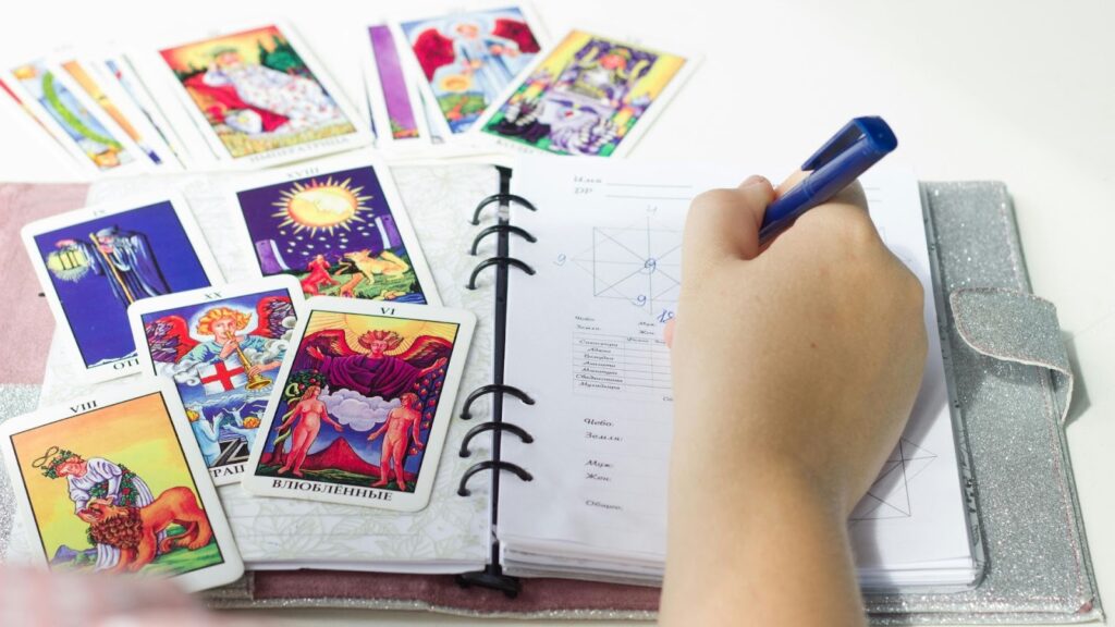  一個人正在筆記本上記錄占卜結果，旁邊散落著多張彩繪塔羅牌，畫面呈現專業研究與紀錄的氛圍。 