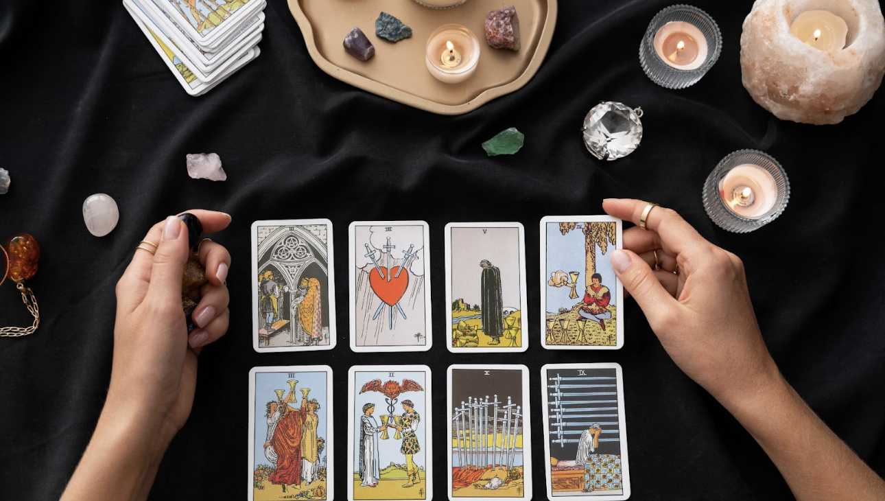 一雙手正在黑色桌面上進行塔羅牌占卜，桌上擺放著多張塔羅牌、水晶原礦、透明球體及數盞點燃的蠟燭