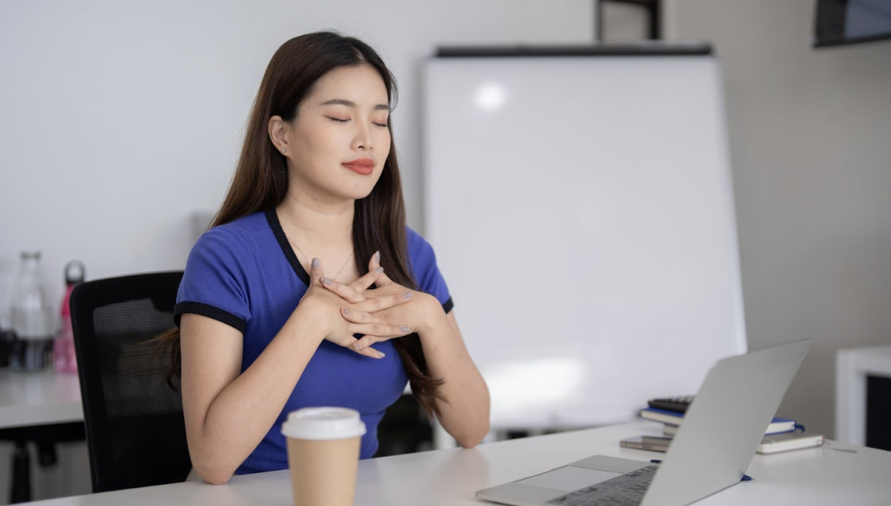 一名身穿藍色上衣的女性坐在辦公桌前，閉上雙手輕撫胸口深呼吸，展現出在忙碌工作中找尋平靜的瞬間