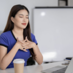 一名身穿藍色上衣的女性坐在辦公桌前，閉上雙手輕撫胸口深呼吸，展現出在忙碌工作中找尋平靜的瞬間