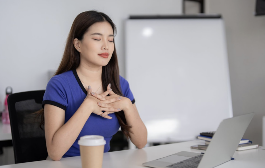 一名身穿藍色上衣的女性坐在辦公桌前，閉上雙手輕撫胸口深呼吸，展現出在忙碌工作中找尋平靜的瞬間