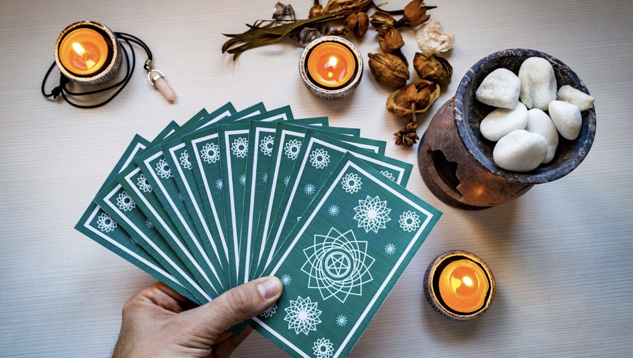 一隻手拿著一疊綠色曼陀羅圖案的塔羅牌呈扇形展開，背景有蠟燭、粉晶與擴香石，營造神祕氛圍