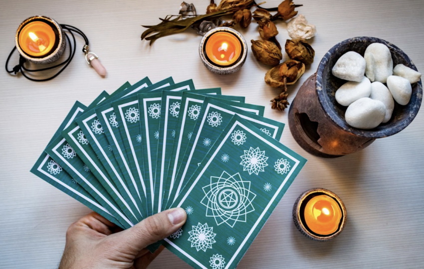一隻手拿著一疊綠色曼陀羅圖案的塔羅牌呈扇形展開，背景有蠟燭、粉晶與擴香石，營造神祕氛圍