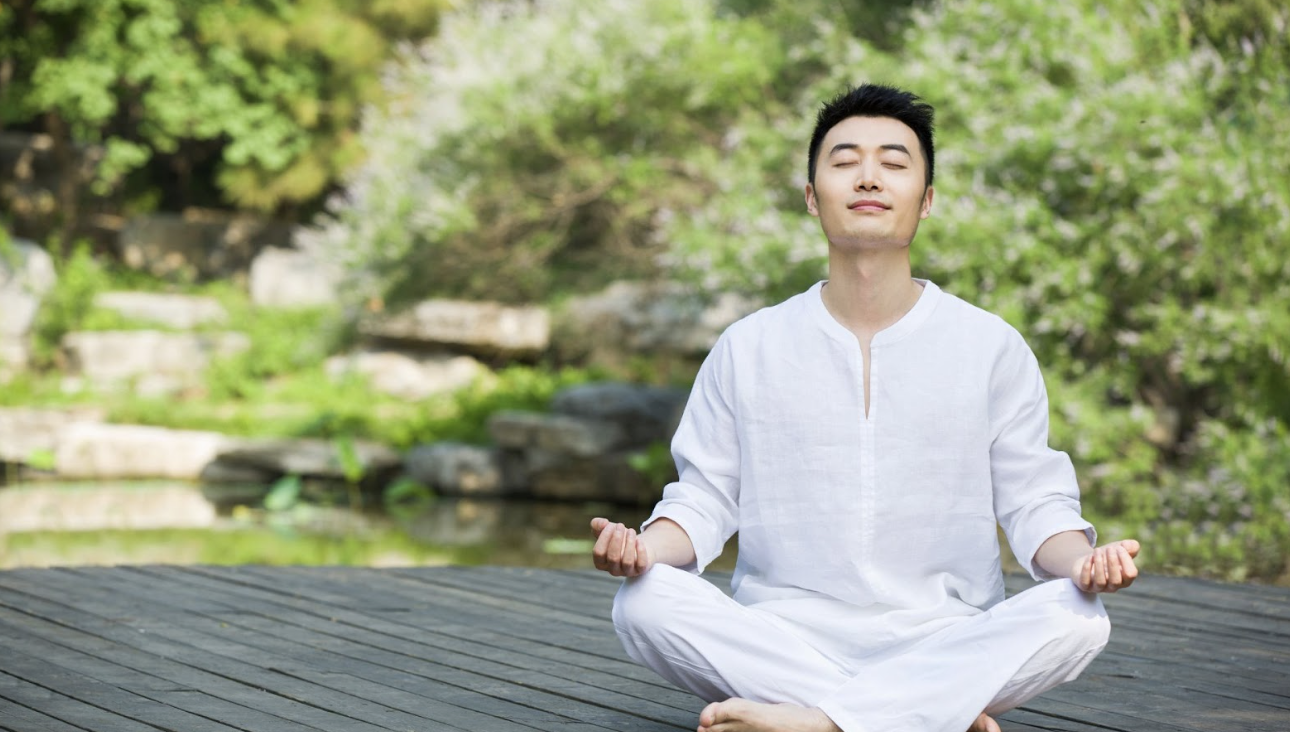 穿著白衣的男子在戶外木棧道上閉目盤坐冥想，背景為綠意盎然的庭園造景。