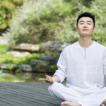 穿著白衣的男子在戶外木棧道上閉目盤坐冥想，背景為綠意盎然的庭園造景。