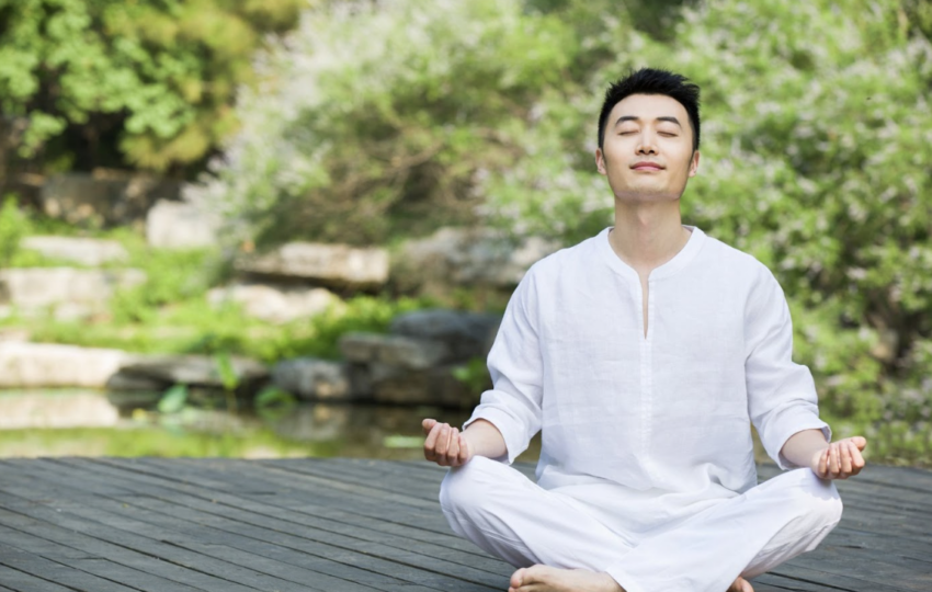 穿著白衣的男子在戶外木棧道上閉目盤坐冥想，背景為綠意盎然的庭園造景。