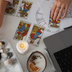 在白色桌面上進行塔羅占卜，畫面上方可見占星命盤圖表與筆記型電腦，占卜師的手正翻動著塔羅牌，周遭擺放著蠟燭、能量原石與香氛裝飾