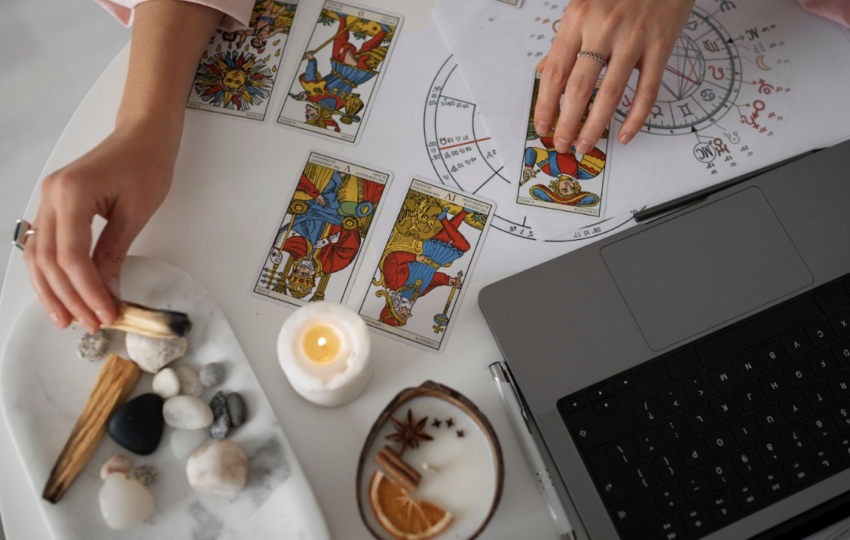 在白色桌面上進行塔羅占卜，畫面上方可見占星命盤圖表與筆記型電腦，占卜師的手正翻動著塔羅牌，周遭擺放著蠟燭、能量原石與香氛裝飾