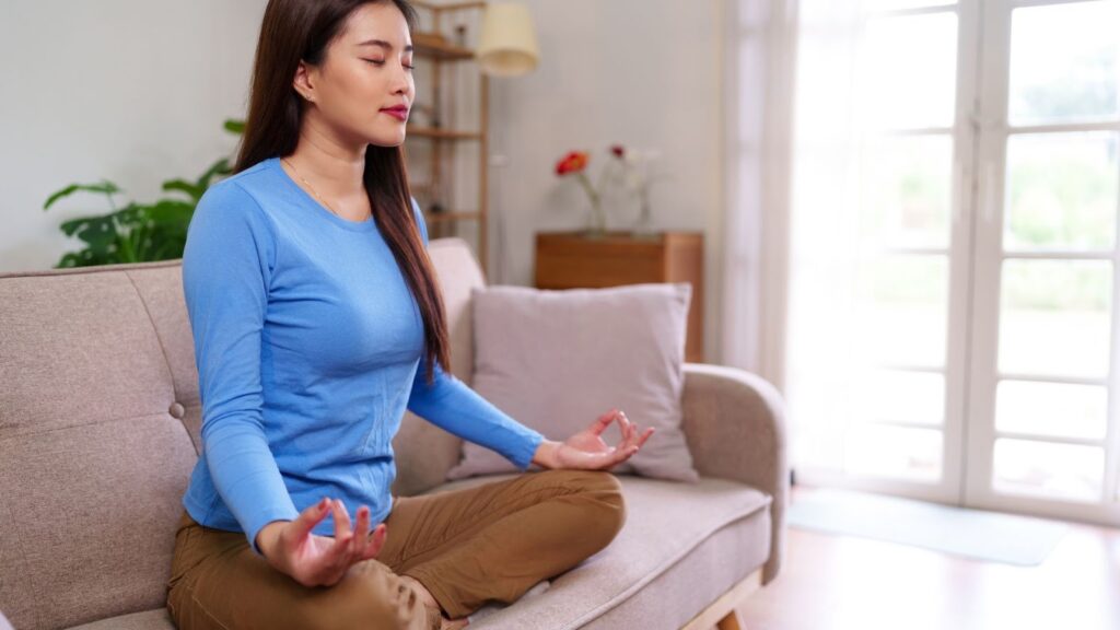 穿著藍色上衣的年輕女性在客廳沙發上盤腿靜坐，表情平和安詳，展現日常生活中的正念練習。