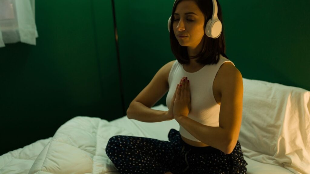 女子在昏暗的臥室戴著耳機專注冥想，雙手合十祈禱] 敘述： 透過冥想音樂與引導音頻，在夜晚打造專屬的靈魂角落。雙手合十，內心默唸感謝，為一天畫下平穩的句點。