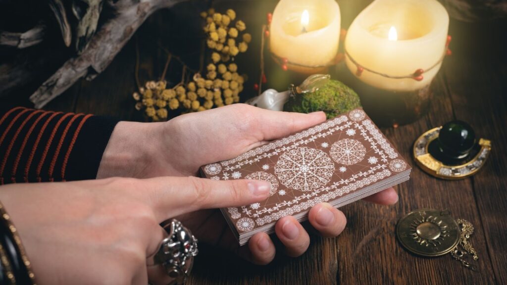 特寫雙手觸摸疊好的棕色幾何花紋塔羅牌，背景有燃燒的蠟燭、乾花與水晶球，營造神祕占卜氛圍