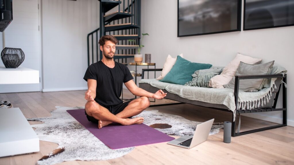 男子在客廳的紫色瑜伽墊上盤腿冥想，前方放著筆電，似乎正參與線上冥想課程或視訊練習。