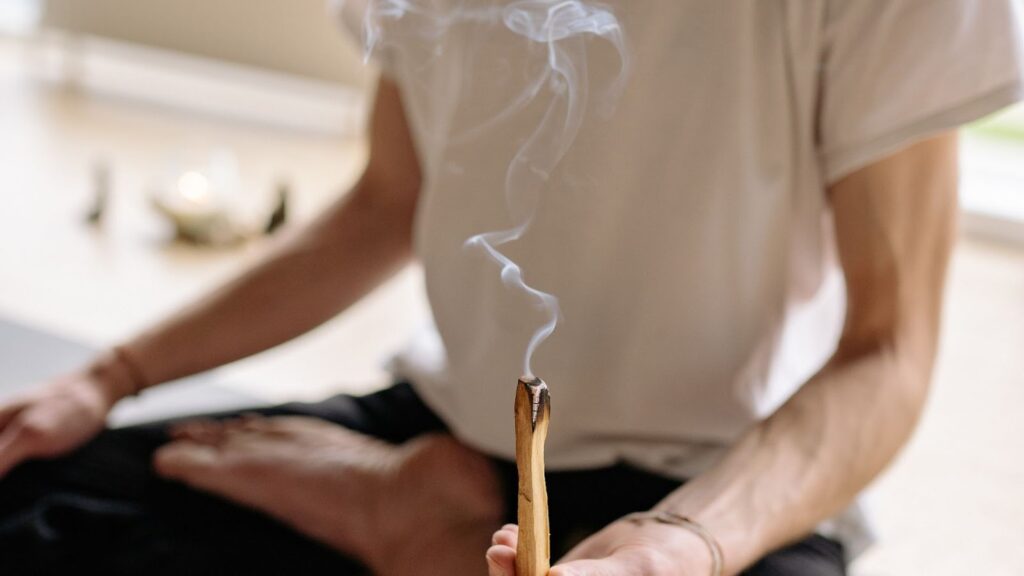 特寫一根正在燃燒並冒出白煙的秘魯聖木（Palo Santo），背景為一名正在靜坐的身影，用於空間淨化