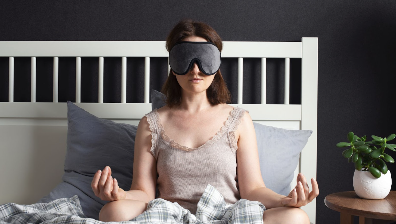 女子戴著灰色睡眠眼罩在床上盤腿坐著進行冥想] 敘述： 遮斷視覺的干擾，能讓感官更往內在聚焦。在睡前進行簡短的冥想，是提升睡眠品質與緩解焦慮的絕佳練習。