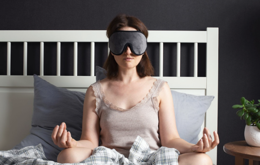 女子戴著灰色睡眠眼罩在床上盤腿坐著進行冥想] 敘述： 遮斷視覺的干擾，能讓感官更往內在聚焦。在睡前進行簡短的冥想，是提升睡眠品質與緩解焦慮的絕佳練習。