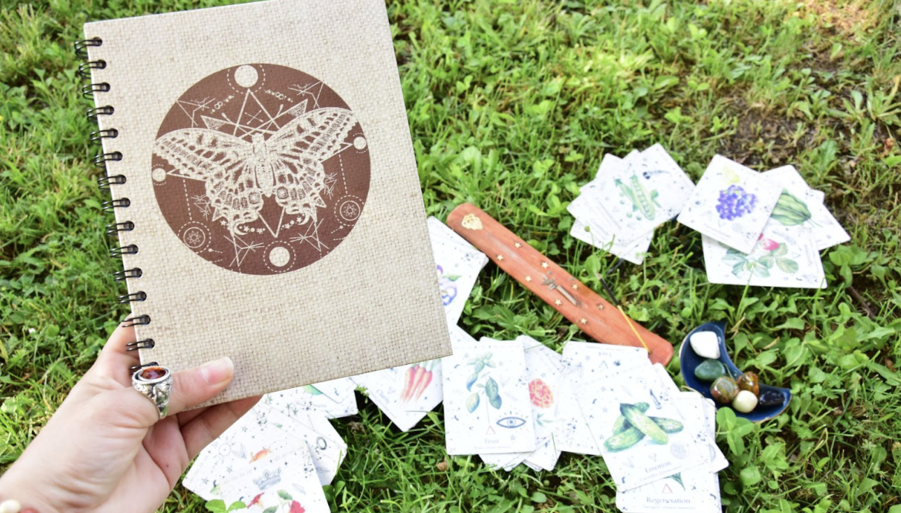 一隻戴著復古銀戒的手拿著一本封面印有蝴蝶幾何圖騰的麻布筆記本，背景是草地上散落的塔羅牌與薰香棒