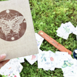 一隻戴著復古銀戒的手拿著一本封面印有蝴蝶幾何圖騰的麻布筆記本，背景是草地上散落的塔羅牌與薰香棒