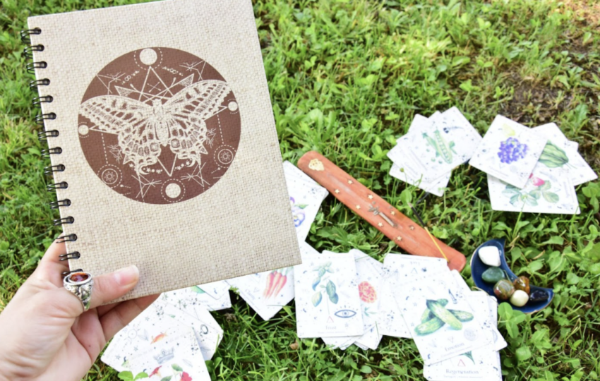 一隻戴著復古銀戒的手拿著一本封面印有蝴蝶幾何圖騰的麻布筆記本，背景是草地上散落的塔羅牌與薰香棒