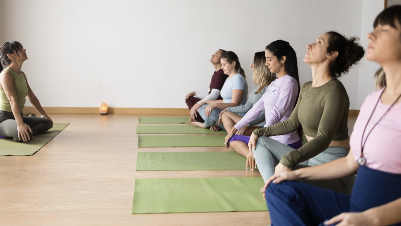 多名學員在明亮的教室內排成一列，閉眼盤腿坐在綠色瑜伽墊上，跟隨前方的老師進行靜坐引導