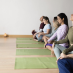 多名學員在明亮的教室內排成一列，閉眼盤腿坐在綠色瑜伽墊上，跟隨前方的老師進行靜坐引導