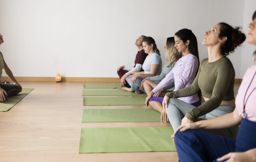 多名學員在明亮的教室內排成一列，閉眼盤腿坐在綠色瑜伽墊上，跟隨前方的老師進行靜坐引導