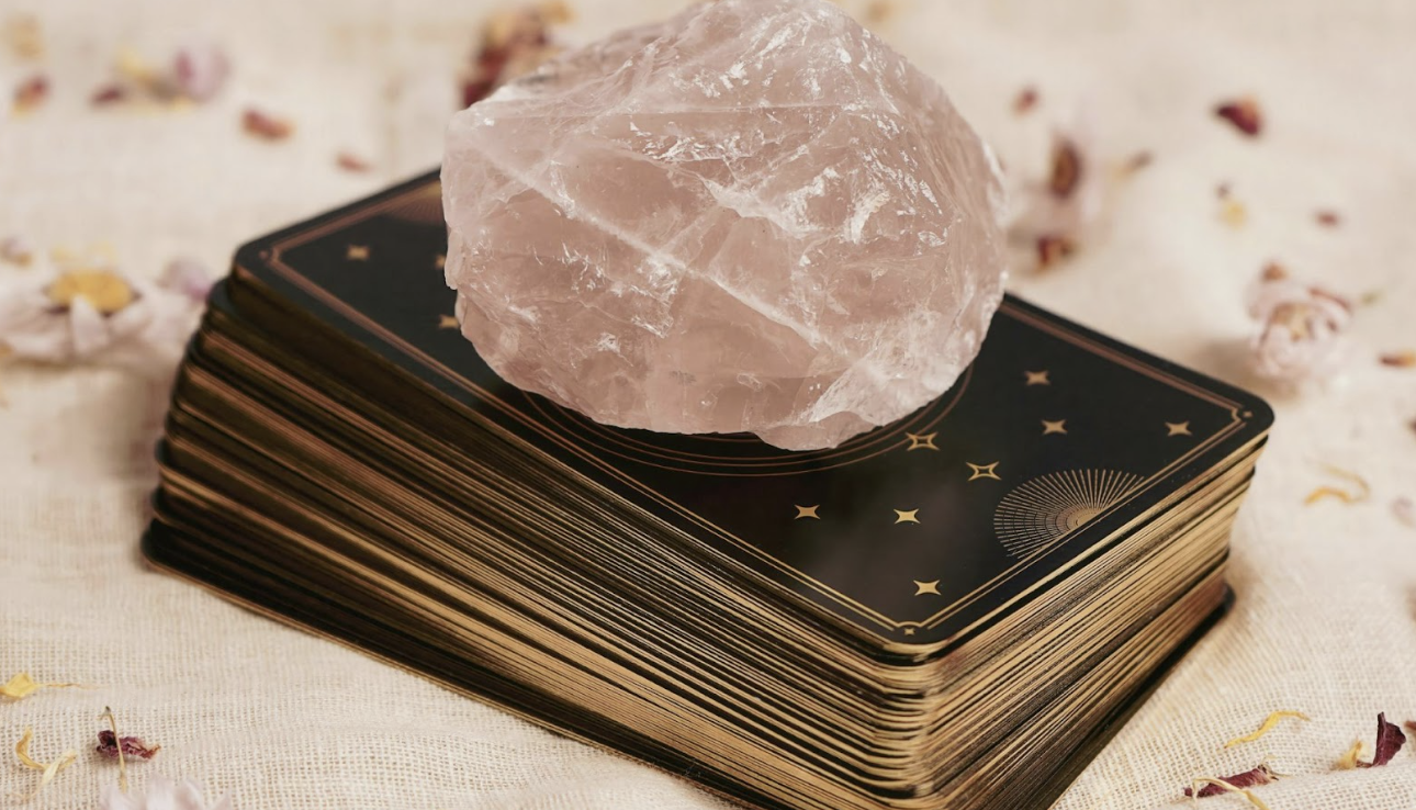 一疊帶有金色星辰圖案的塔羅牌背面，上方放著一顆粉紅色的粉晶原石，用於身心靈能量療癒與占卜