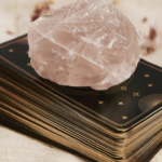 一疊帶有金色星辰圖案的塔羅牌背面，上方放著一顆粉紅色的粉晶原石，用於身心靈能量療癒與占卜