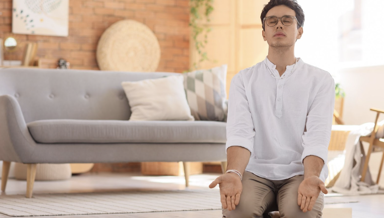 一名男子在客廳溫馨的環境中盤腿坐著進行冥想練習，雙手掌心向上，展現平靜與放鬆的身心狀態