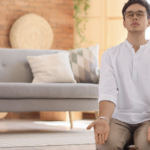一名男子在客廳溫馨的環境中盤腿坐著進行冥想練習，雙手掌心向上，展現平靜與放鬆的身心狀態