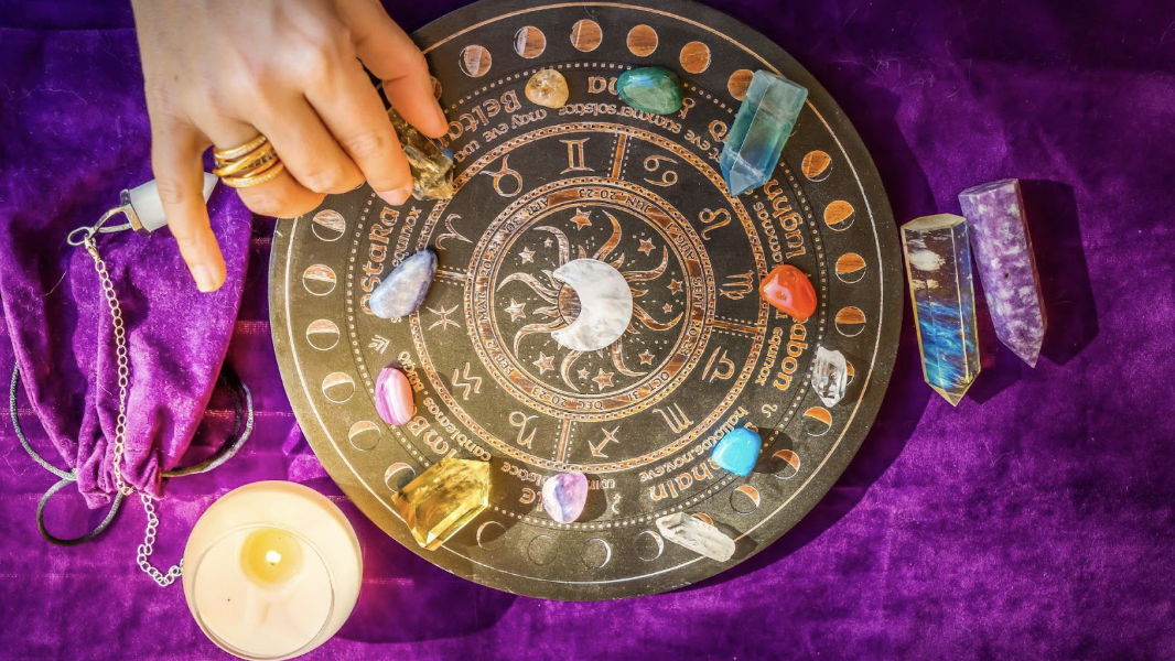 俯瞰圓形占星盤，上面放有多種彩色水晶柱和拋光寶石，一隻戴著戒指的手正在擺放水晶