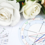白色與淡橘色玫瑰花朵擺放在占星命盤紙張上，盤面上標示著星座符號與行星相位曲線，呈現優雅神祕的占星學氛圍
