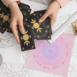 俯瞰占卜師的雙手正在整理帶有月亮週期圖案的黑色塔羅牌，桌面擺放著星座圖表、白色蠟燭、水晶簇以及乾燥植物裝飾